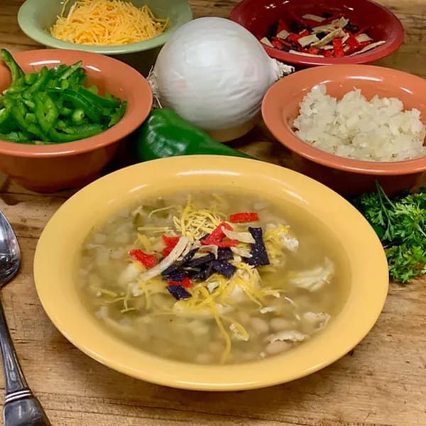 A bowl of Lola's Chicken Shack white chicken chili with toppings and ingredients on a wooden table.