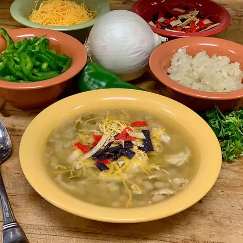 A bowl of Lola's Chicken Shack white chicken chili with toppings and ingredients on a wooden table.