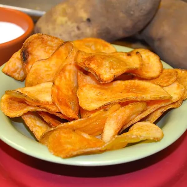 A light green plate filled with crispy, golden-brown potato chips, with a small bowl of dip and raw potatoes in the background.